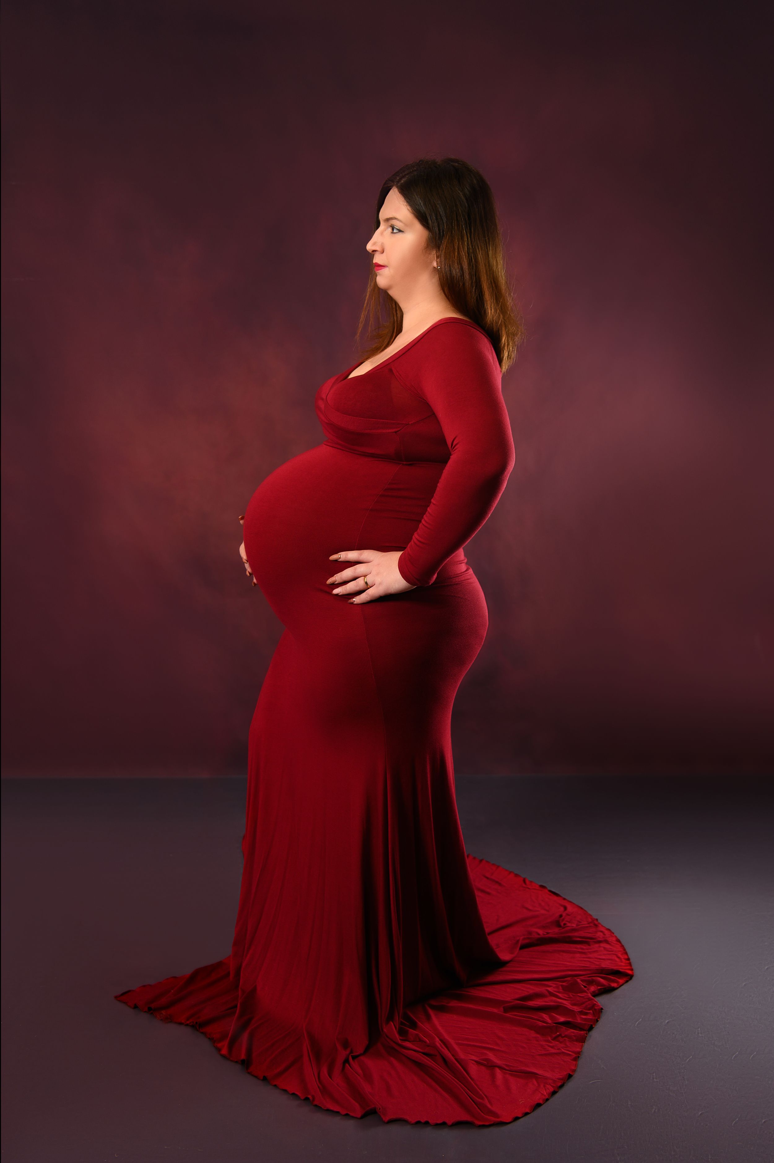 Maternity portrait in flowing red dress against dark dramatic background.