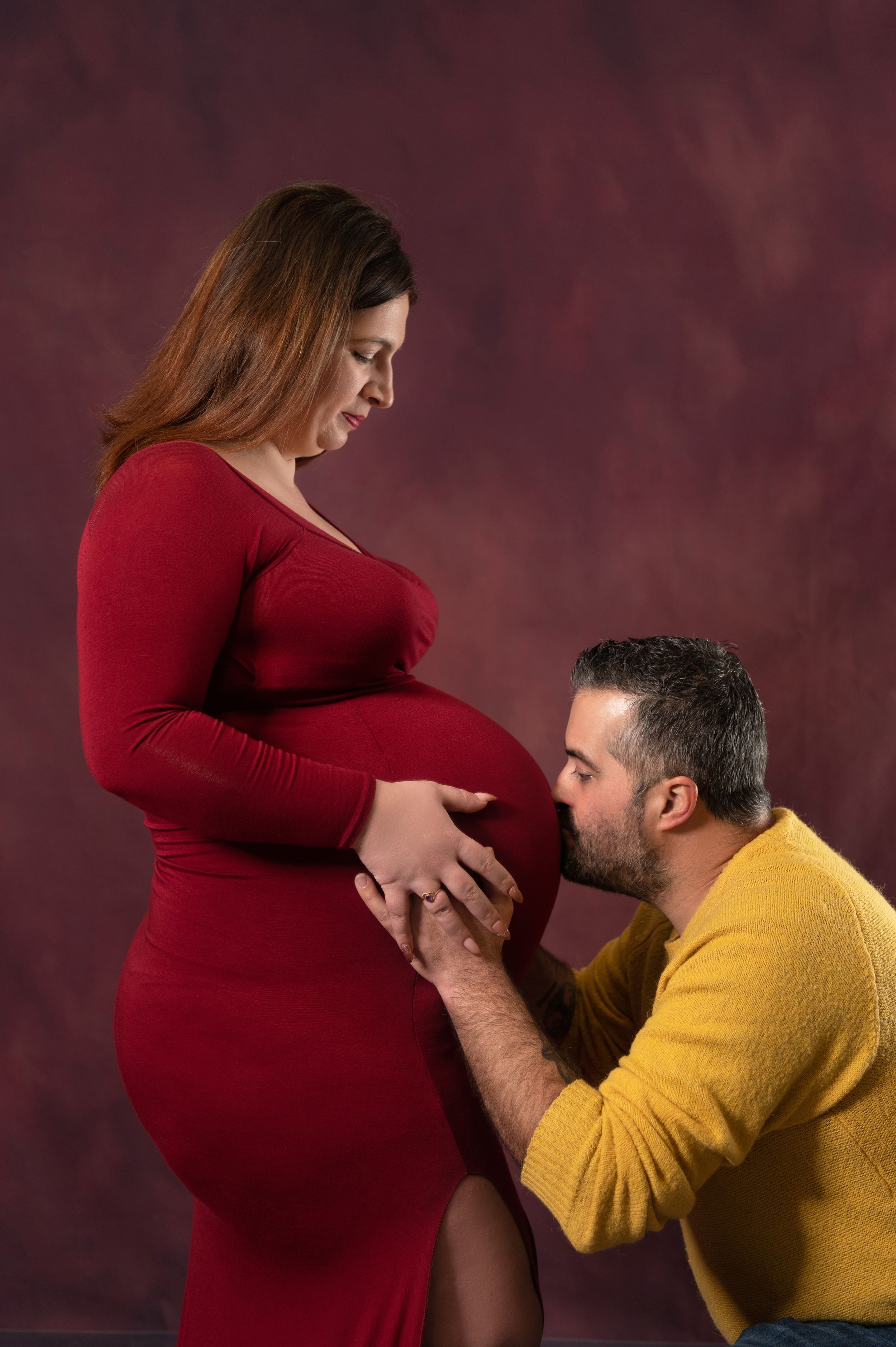 Intimate maternity moment captured against burgundy backdrop.