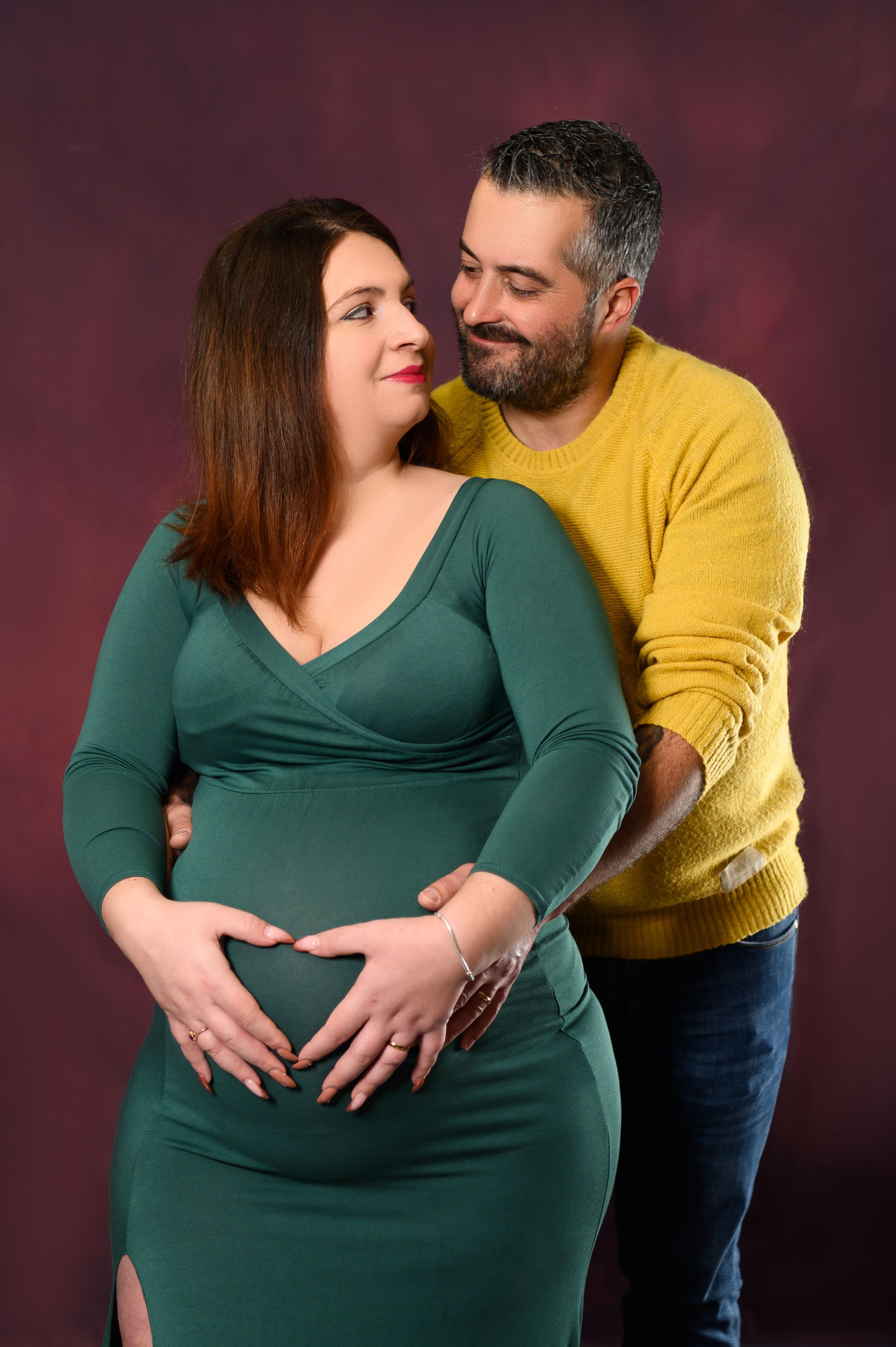 Loving maternity portrait with couple in green and yellow sweaters against dark background.