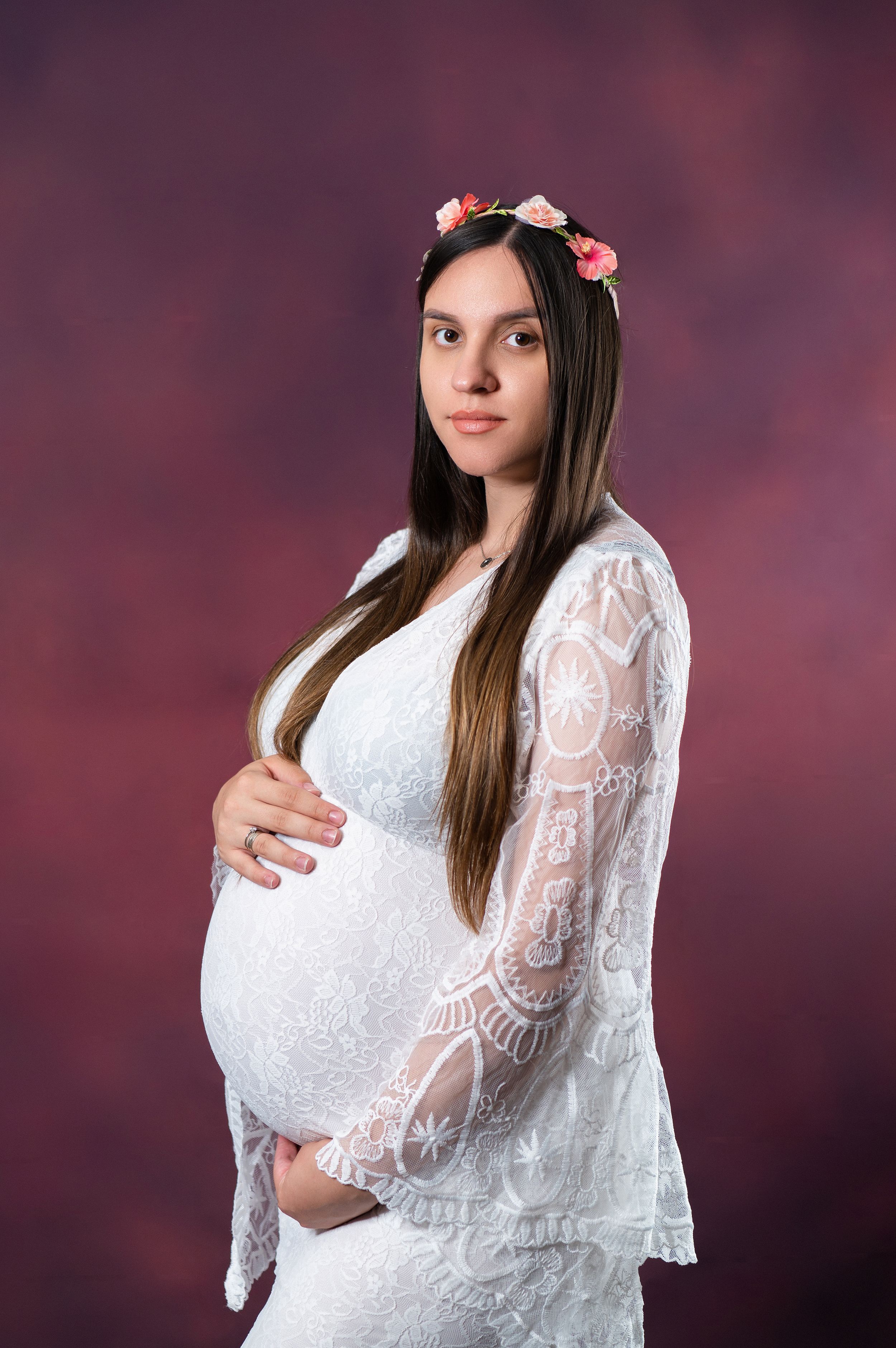 Maternity portrait in white lace dress with bell sleeves against purple backdrop.