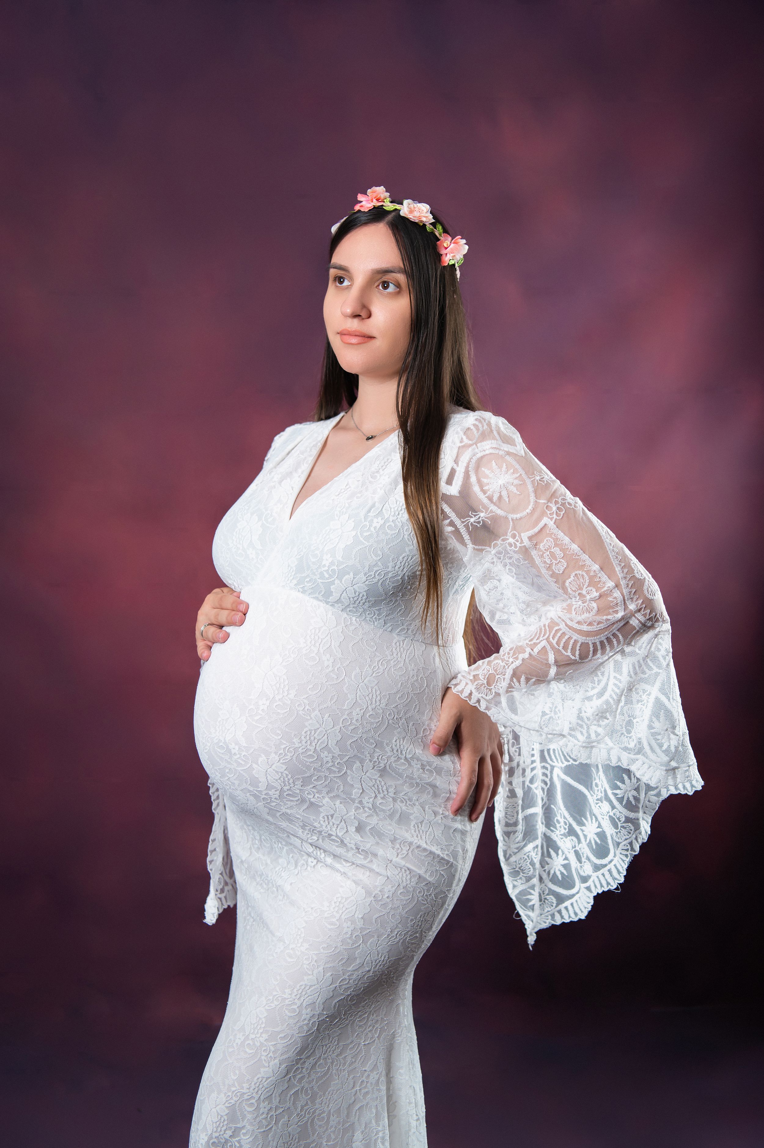 Dramatic maternity pose in white lace gown with bell sleeves against dark backdrop.