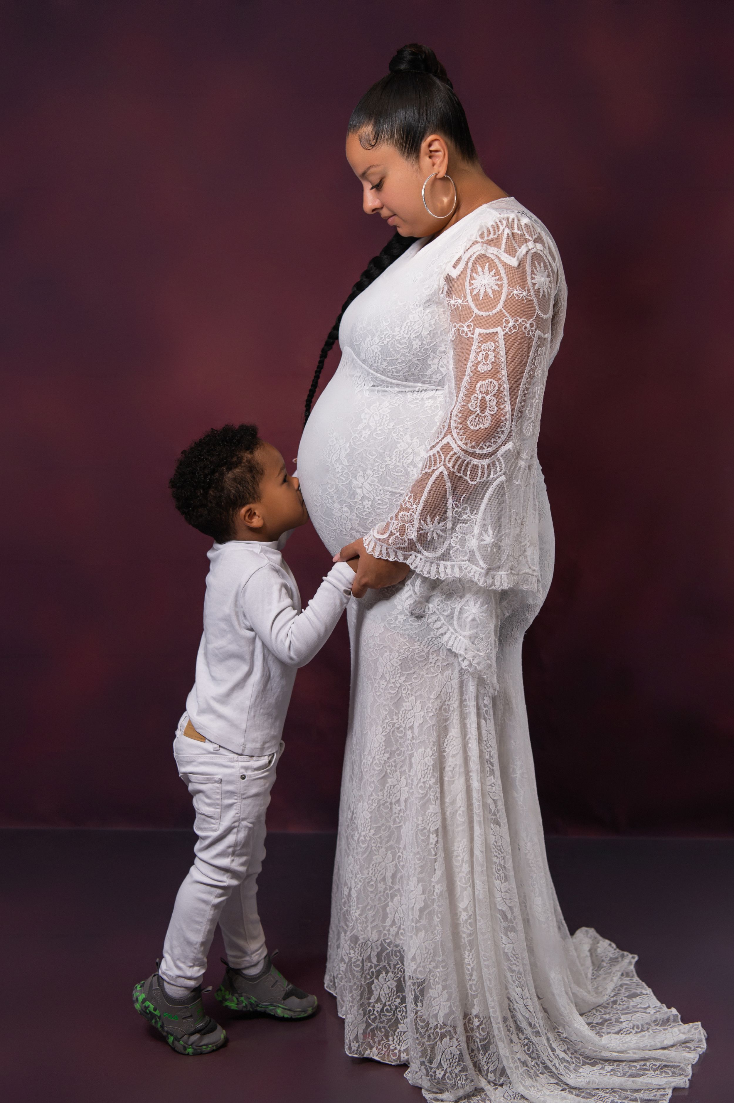 Maternity photoshoot featuring a white lace dress and young child in tender poses against dark background.