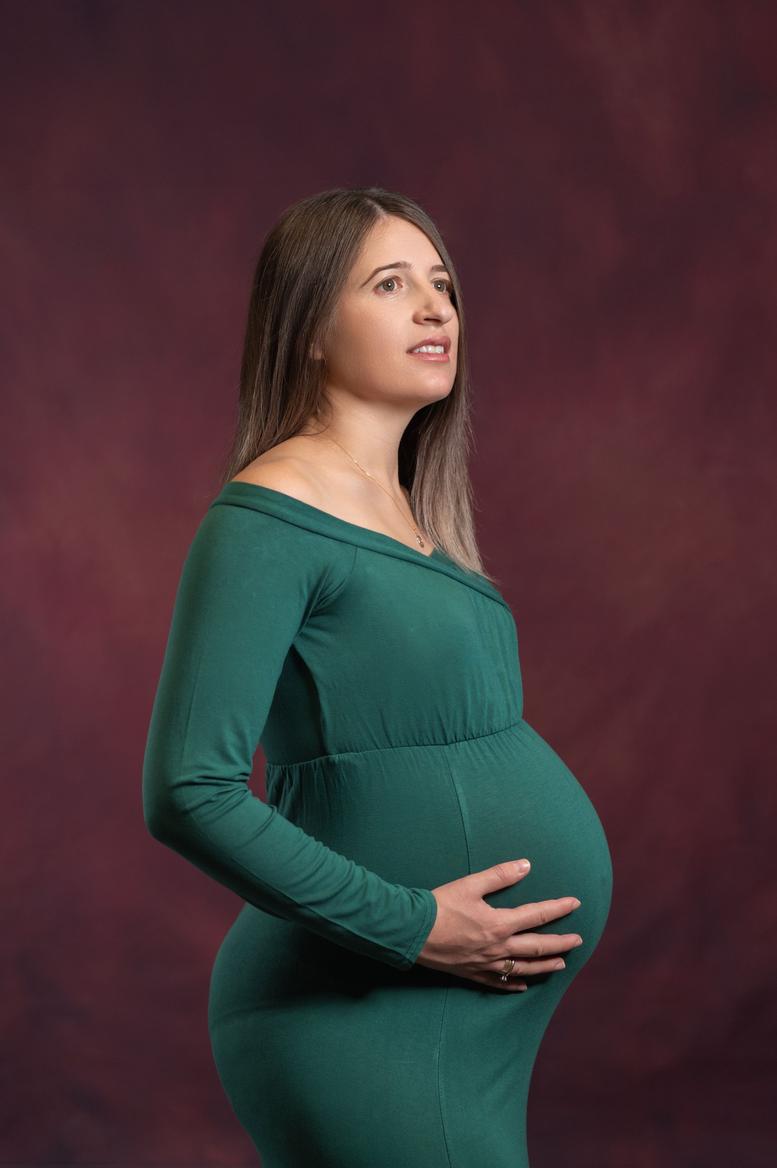 Maternity portraits showing woman in green dress cradling her belly against dark backdrop.