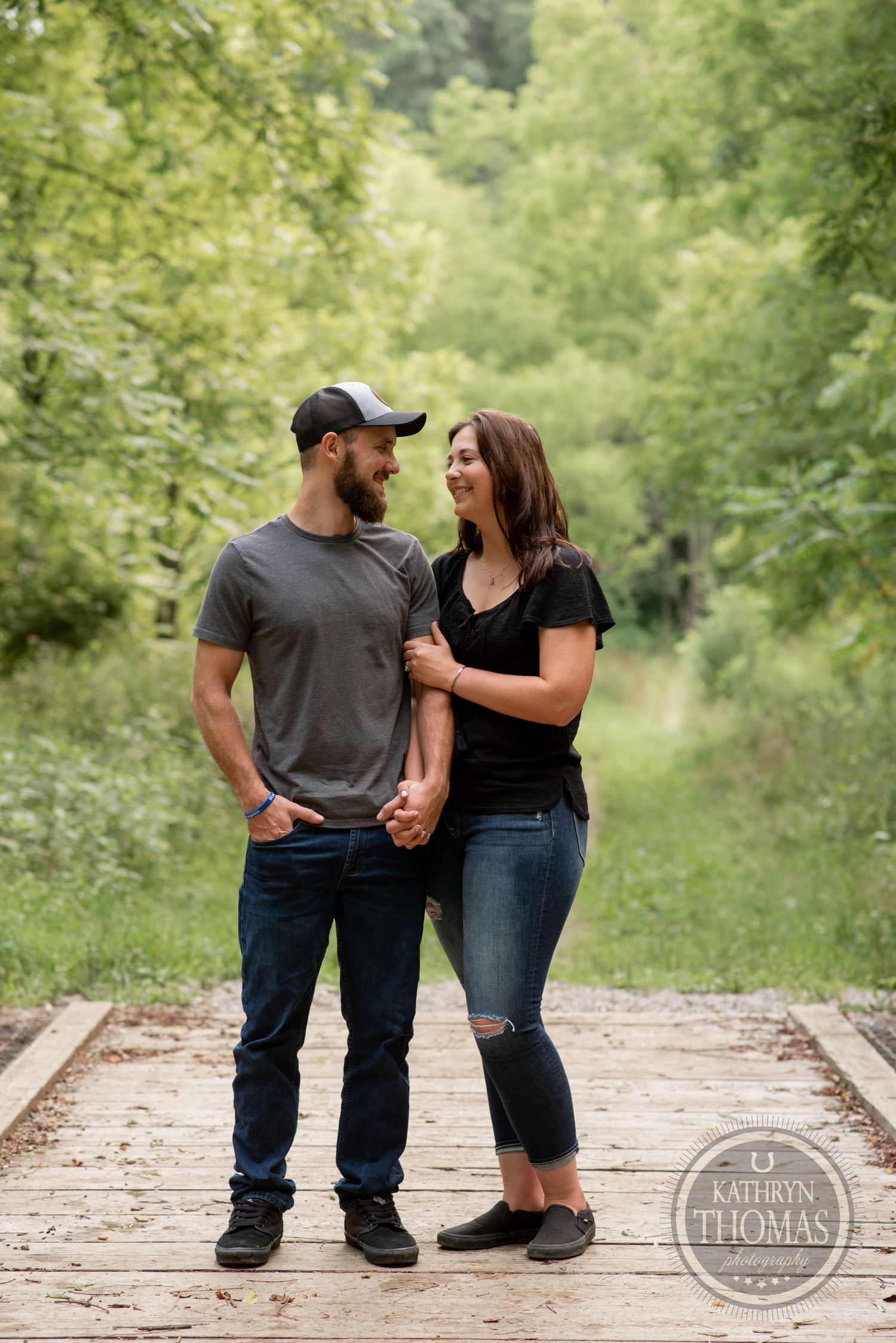 A couple stands close together on wooden railroad tracks in a green forest setting.