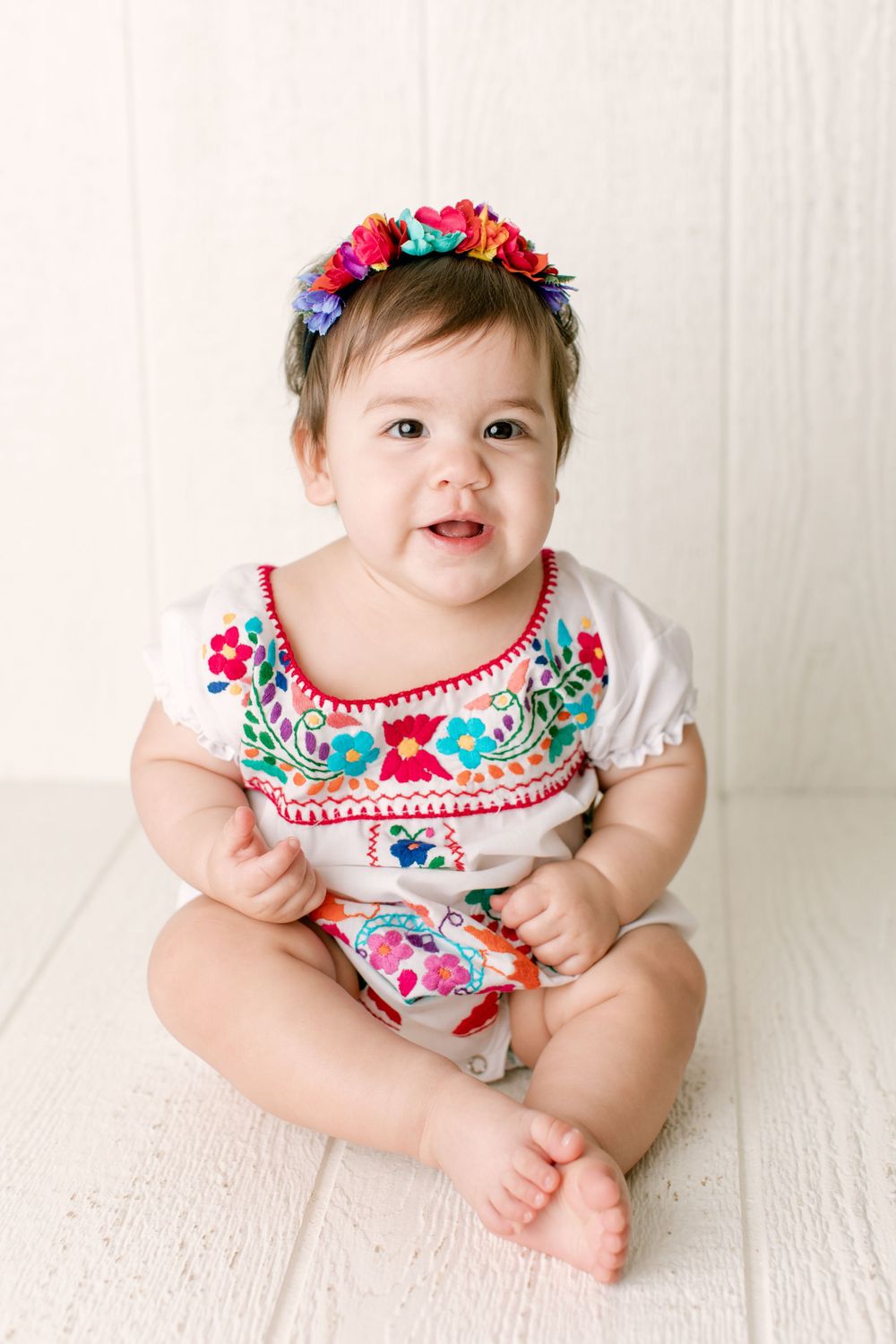 Infant wearing floral embroidered white romper and flower crown sitting on white floor.