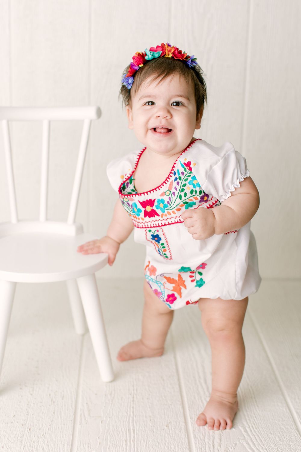 Toddler in embroidered outfit standing next to white chair against white background.