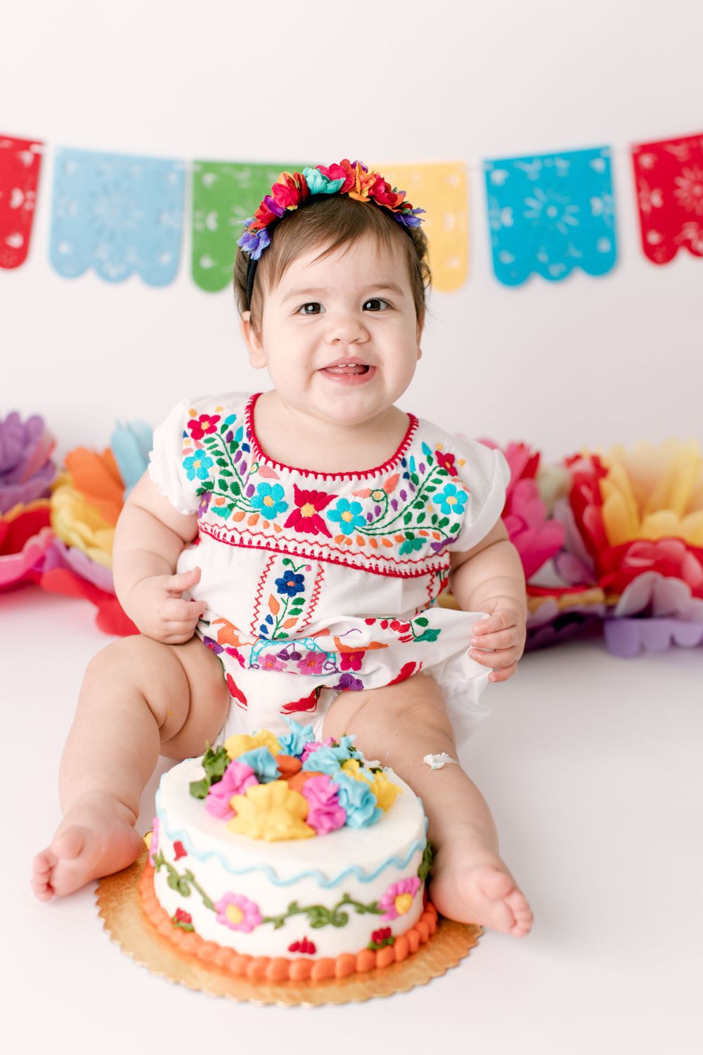 Baby in embroidered outfit with birthday cake under colorful papel picado banners.