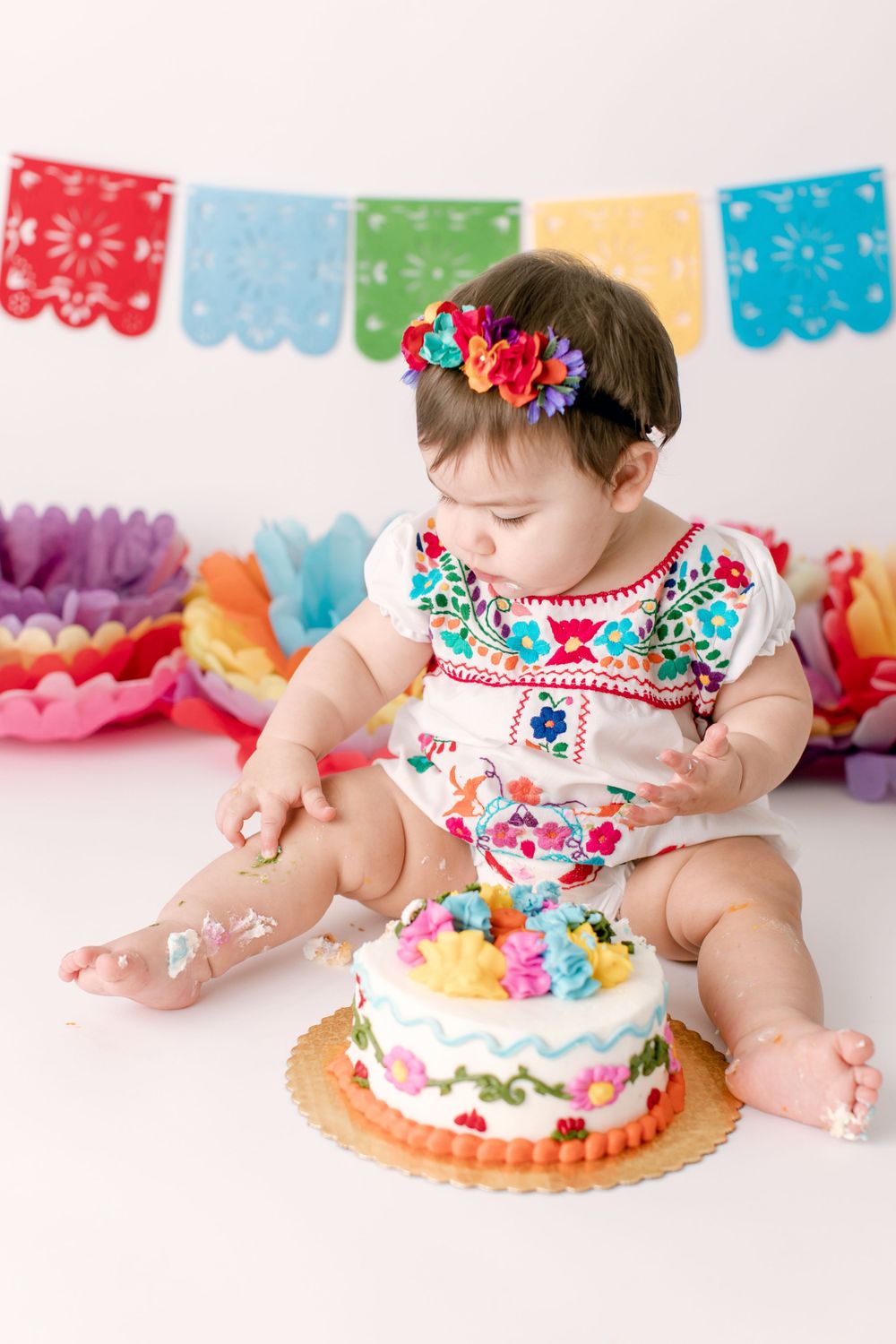 Baby in floral outfit looking down at colorful birthday cake with party decorations.