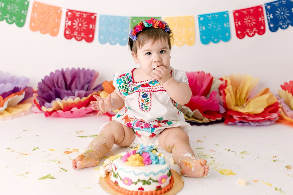 Baby in Mexican-inspired outfit tasting birthday cake with colorful party decorations.