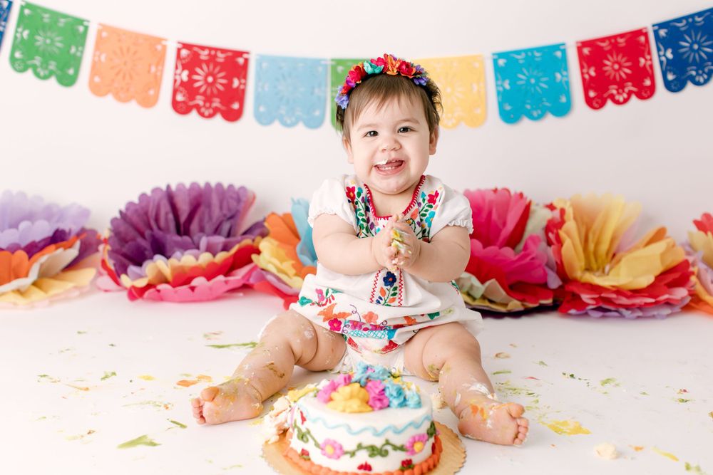 Happy baby in embroidered outfit with birthday cake and colorful fiesta decorations.