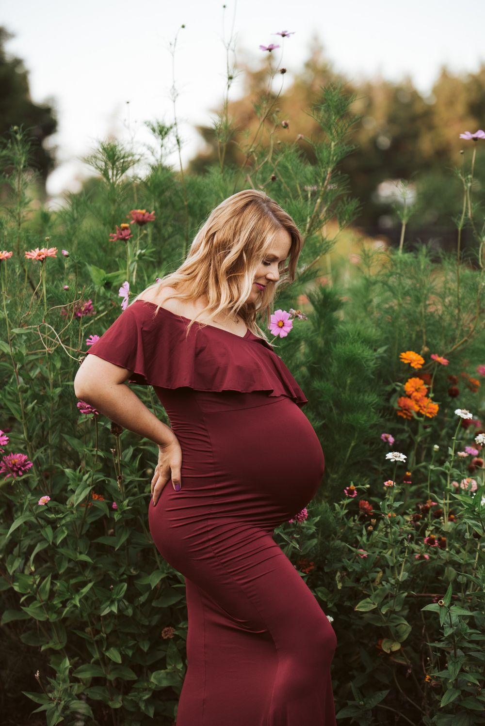 Pregnant woman in burgundy off-shoulder dress standing among colorful wildflowers in garden.