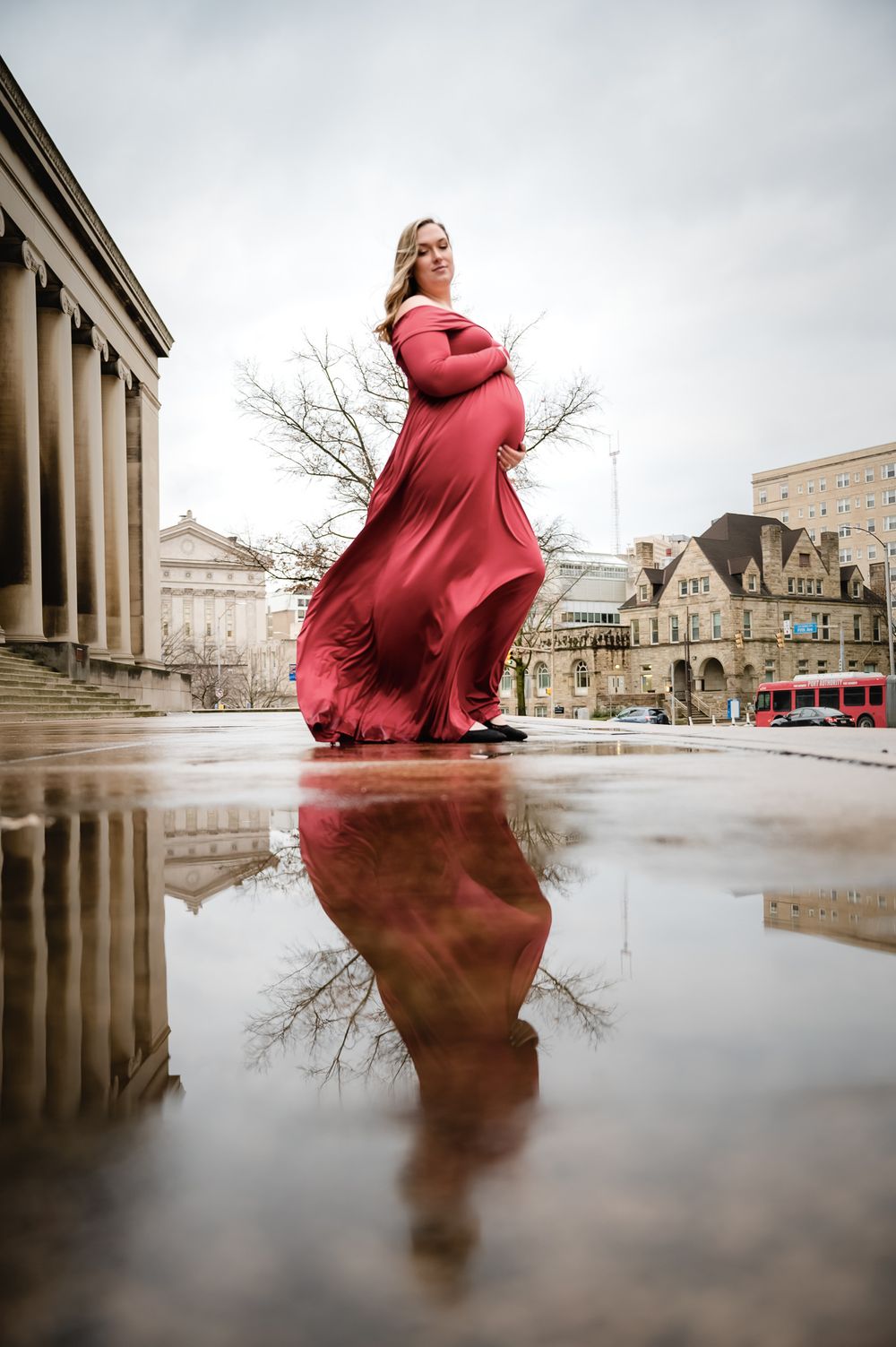 Pregnant woman in flowing red gown reflected in puddle outside historic building.