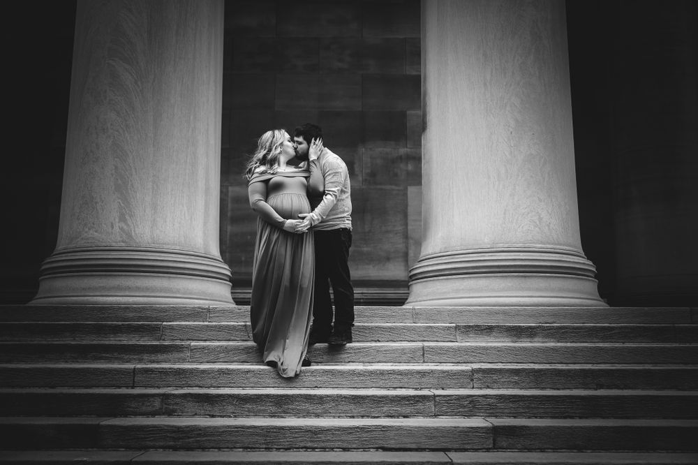Black and white maternity photo of couple embracing on stone steps between grand columns.