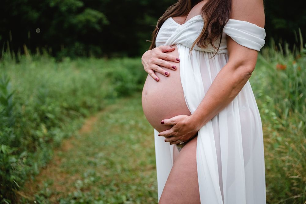 Pregnant woman in white off-shoulder dress cradling bare baby bump in green field.