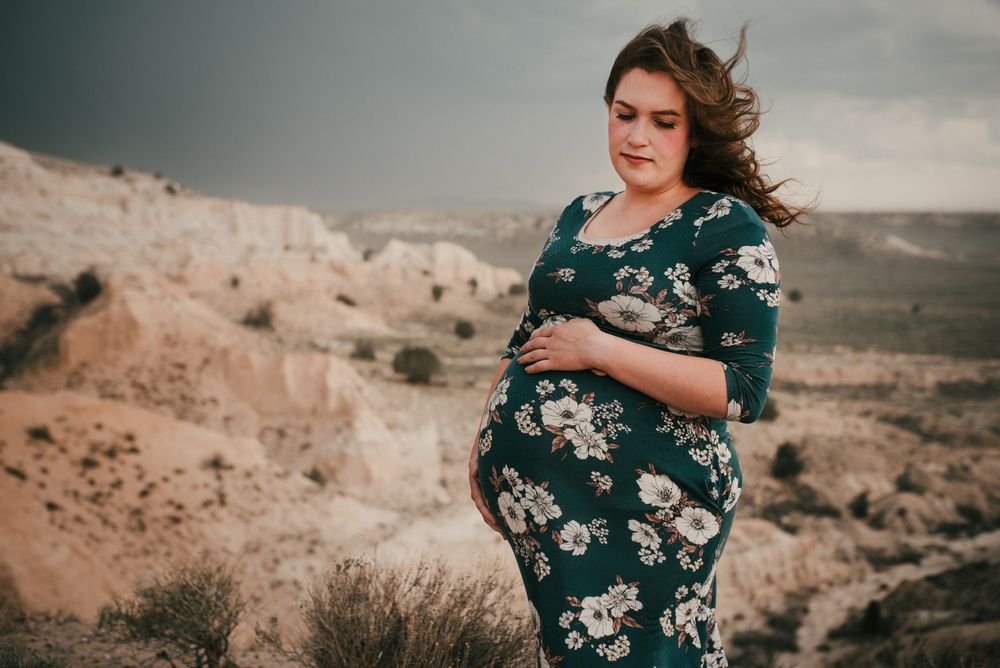 Pregnant woman in floral green dress standing in scenic desert landscape under dramatic sky.