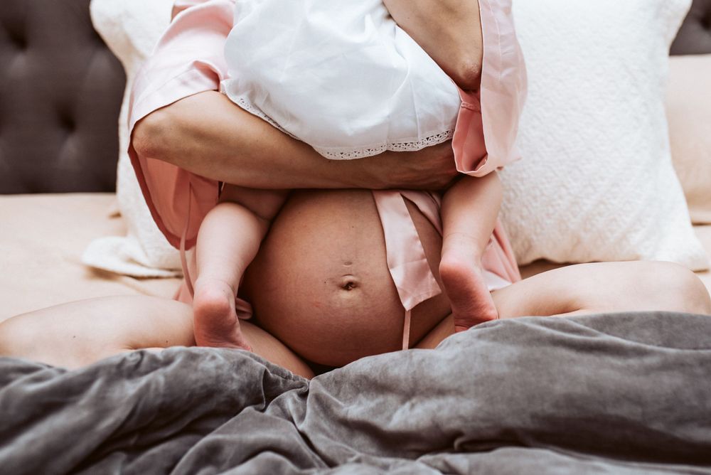 Close-up of pregnant belly with baby sitting on mothers lap in cozy bedroom setting.