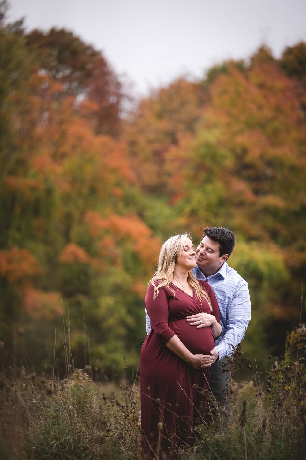 Couple sharing tender moment in autumn forest, man embracing pregnant woman in burgundy dress.