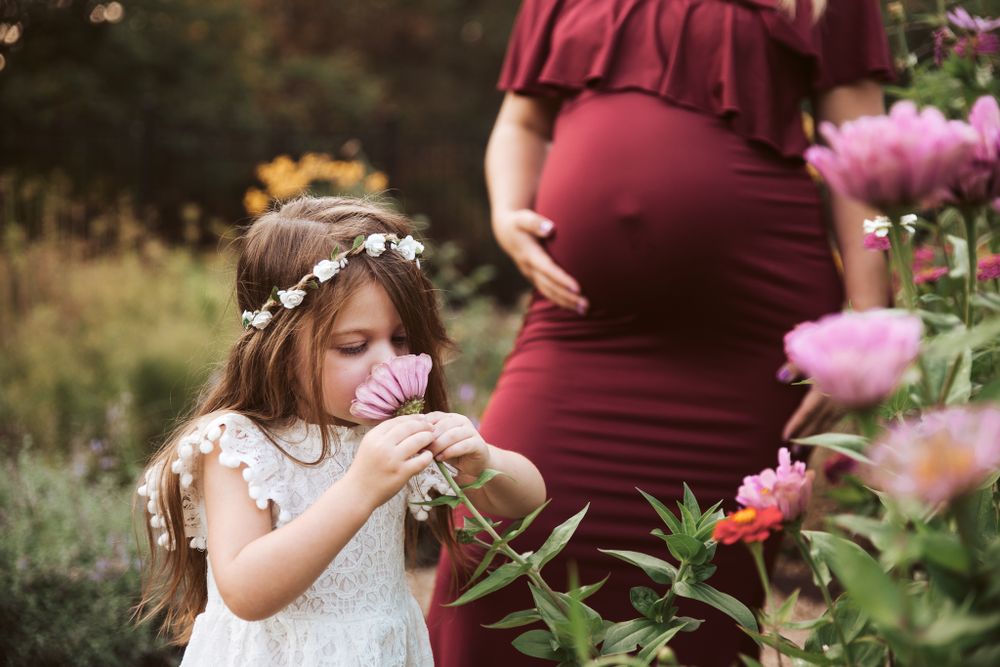 Young girl in floral crown smelling pink flower beside pregnant woman in burgundy dress.