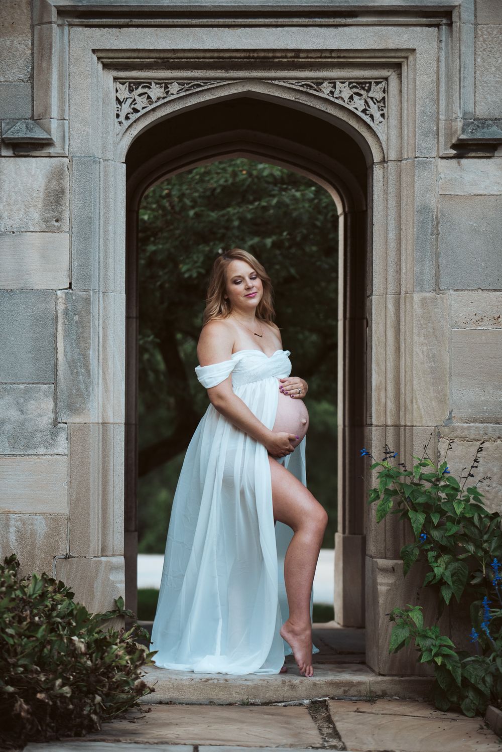 Pregnant woman in sheer white gown posing in ornate gothic stone doorway archway.
