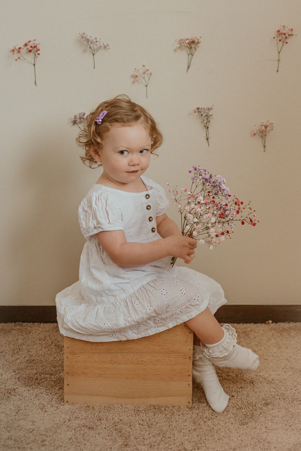 Little girl in white dress holding pink flowers while sitting on wooden box.