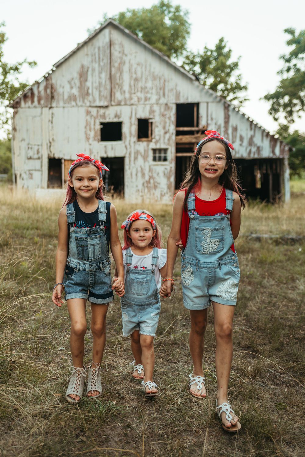 Three kids in denim overalls pose in front of weathered white barn.