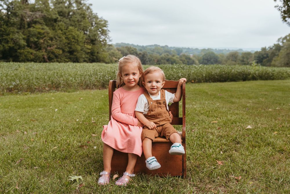 Children sit together on brown wooden bench in sunny meadow.