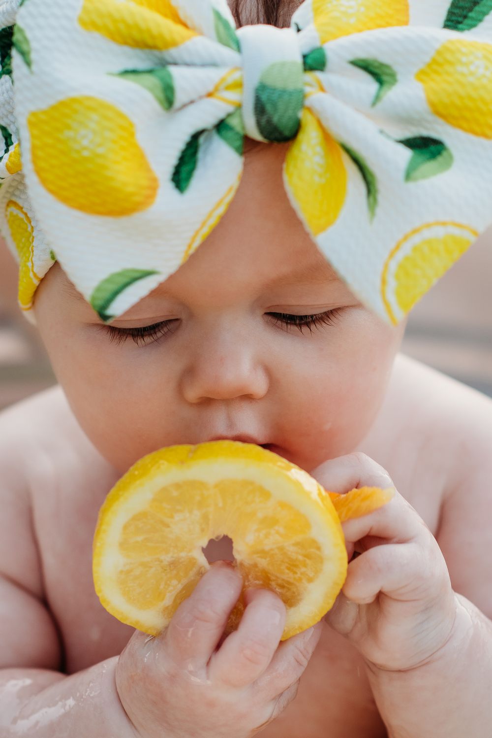 Close up of baby tasting an orange slice while wearing a lemon print bow.