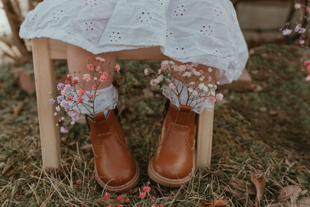 Brown leather boots peek out under a white eyelet dress with dried flowers scattered on the ground.