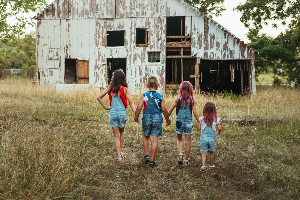 Children holding hands walk toward old white barn in field.