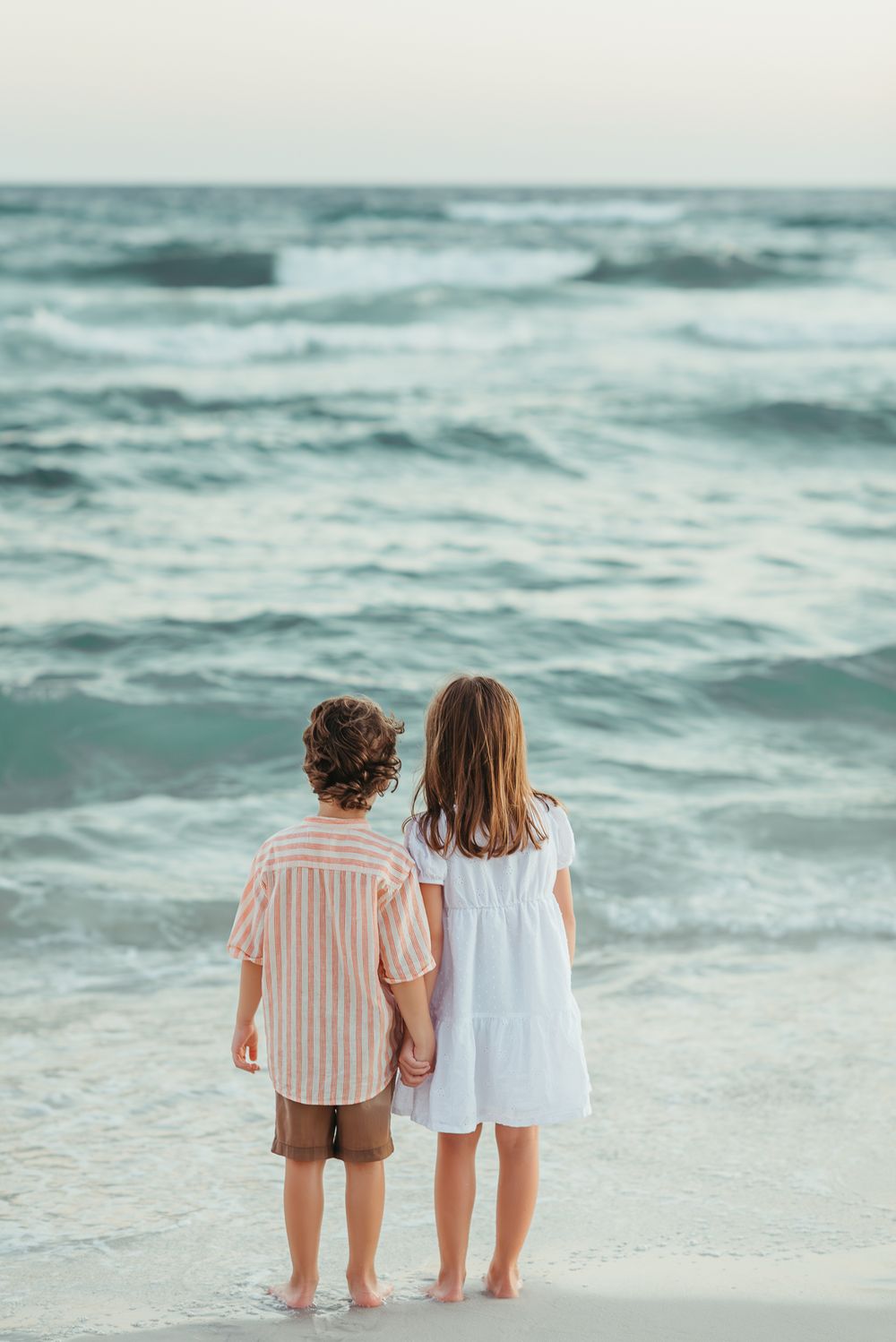 Two small children standing together at the edge of the ocean waves at sunset.