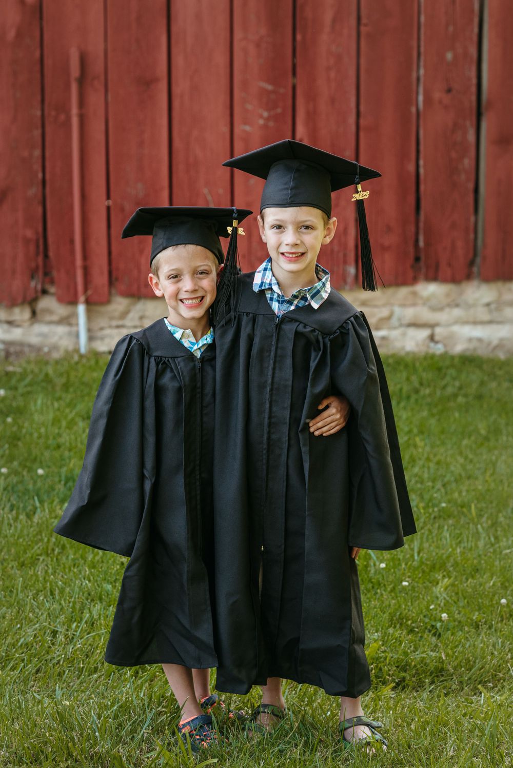 Young graduates in black caps and gowns standing in front of a red barn.