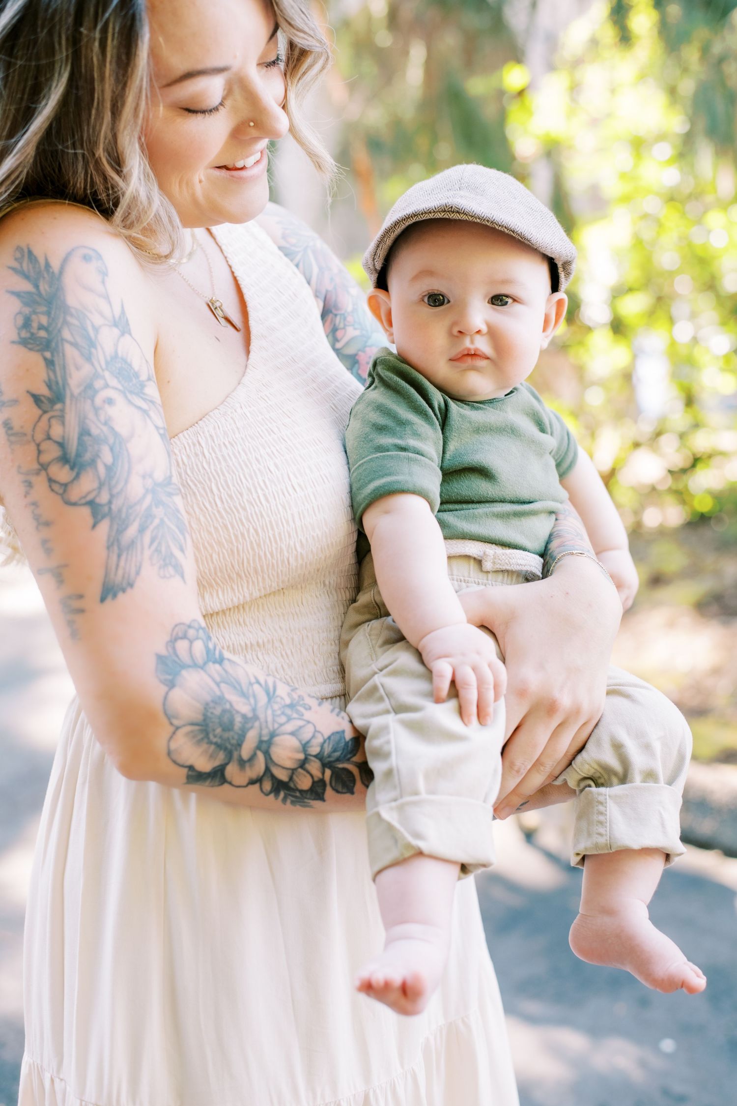 A baby wearing a cap and green shirt is held in white dress against a soft natural background.