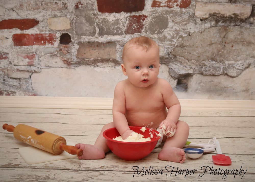Baby sitting on wooden floor with red bowl and rolling pin against brick wall.