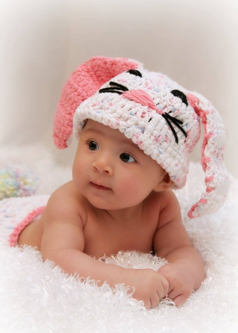 Baby wearing pink and white crochet bunny hat lying on soft white blanket.