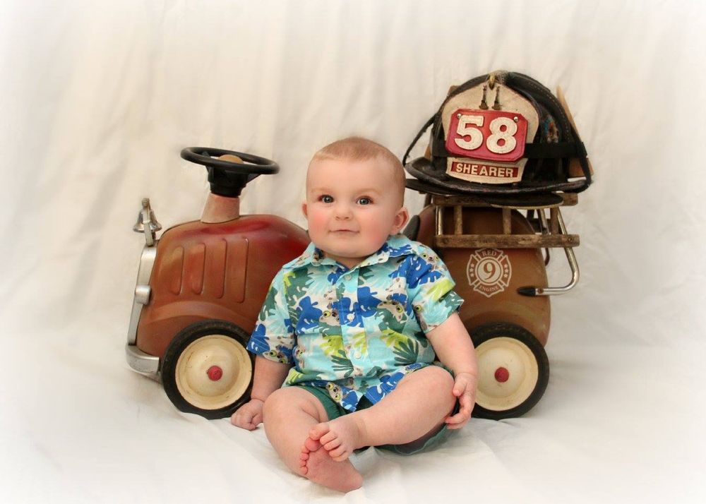 Baby in Hawaiian shirt sitting next to vintage toy wagon and firefighter helmet.