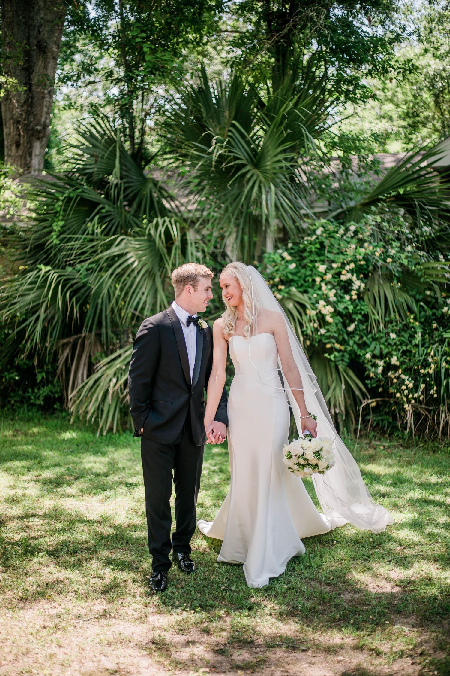 Bride in elegant white gown and groom in tuxedo walk together through a lush garden setting.