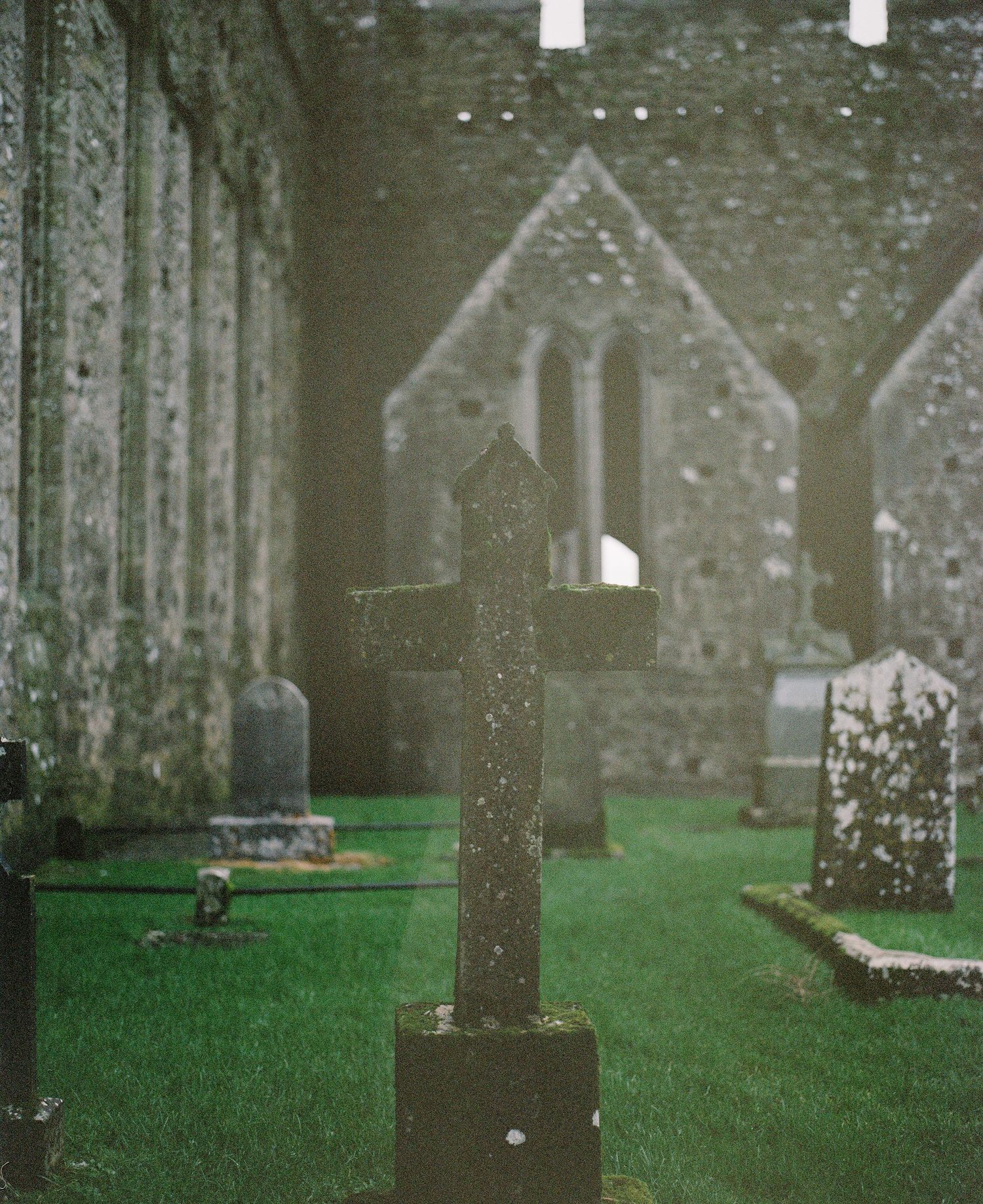 Ancient stone cross and gravestones stand in a misty churchyard with gothic architecture in the background.