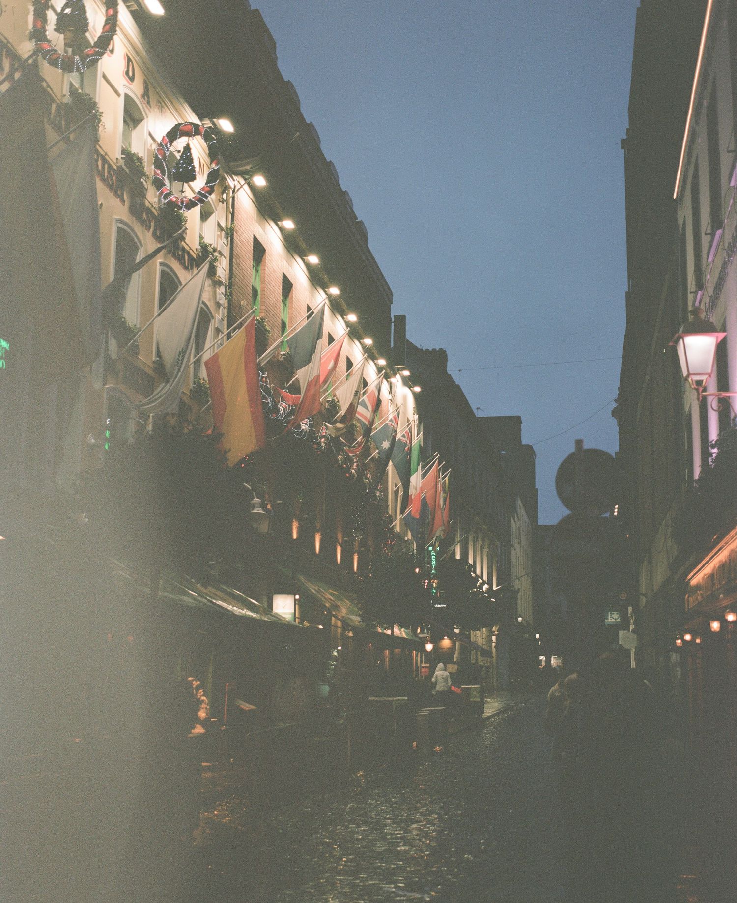 Dark city street with illuminated buildings and strings of lights creating an atmospheric nighttime scene.