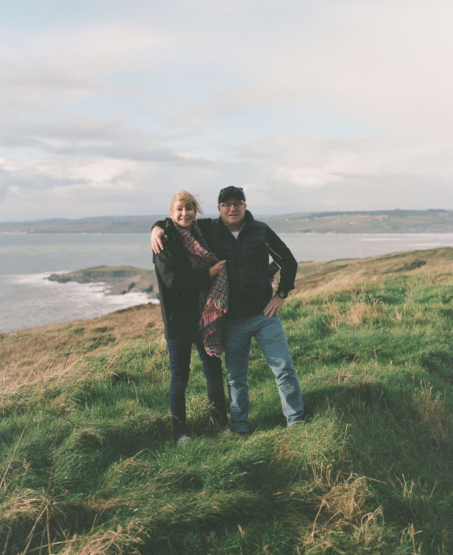 Two people standing on a coastal cliff with ocean views in the background.