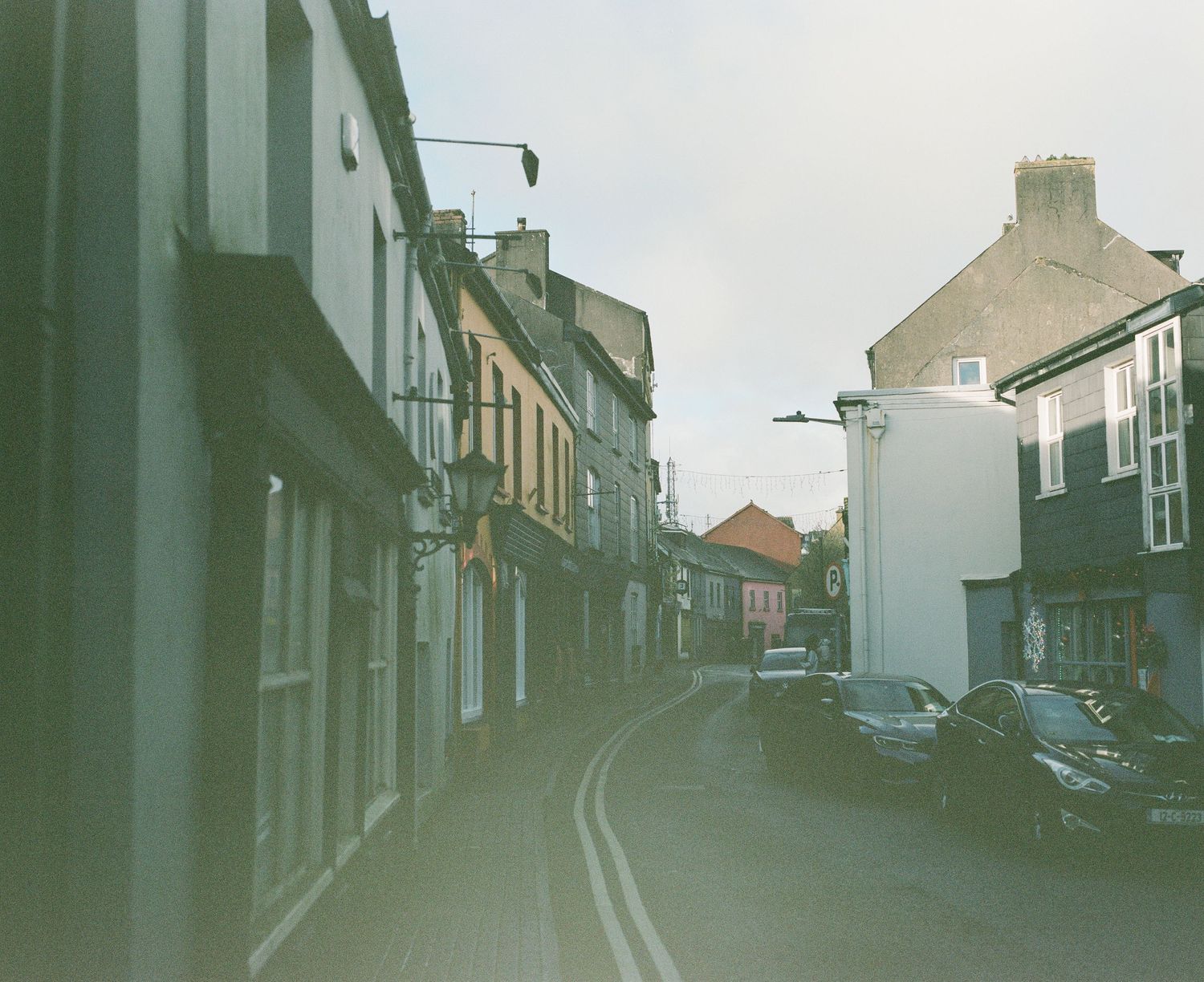 Narrow cobblestone street lined with traditional buildings and parked cars in a small town.