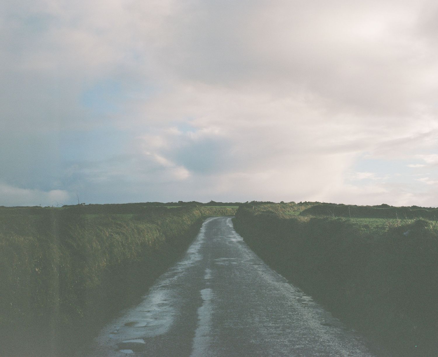Winding dirt road through green fields under cloudy skies.