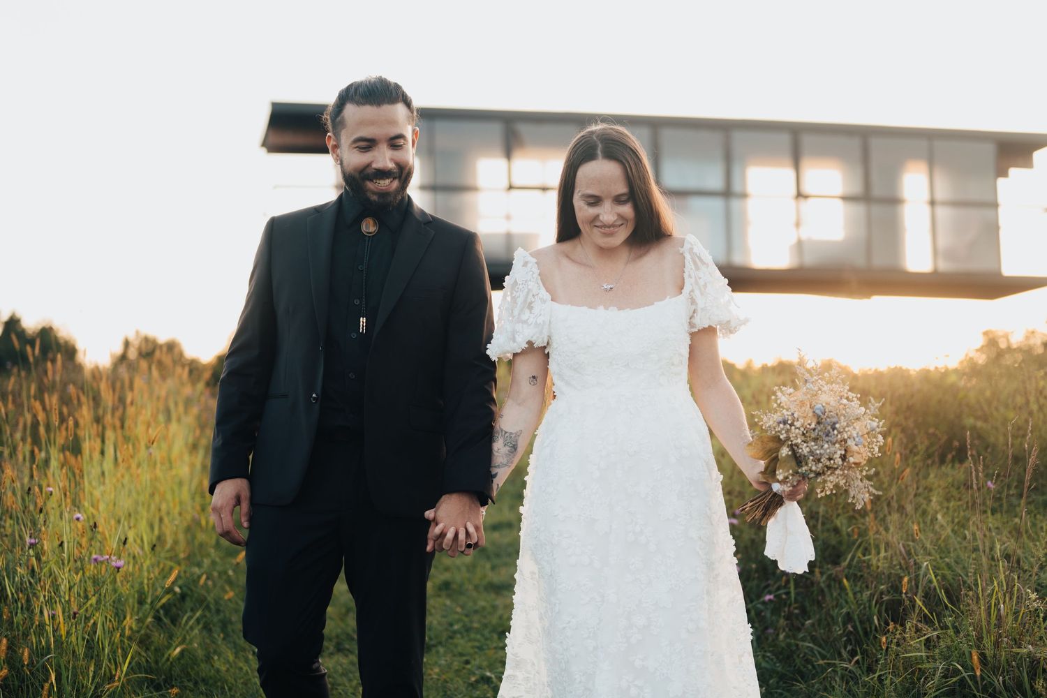 Bride and groom walking hand in hand through a sunny meadow on their wedding day.