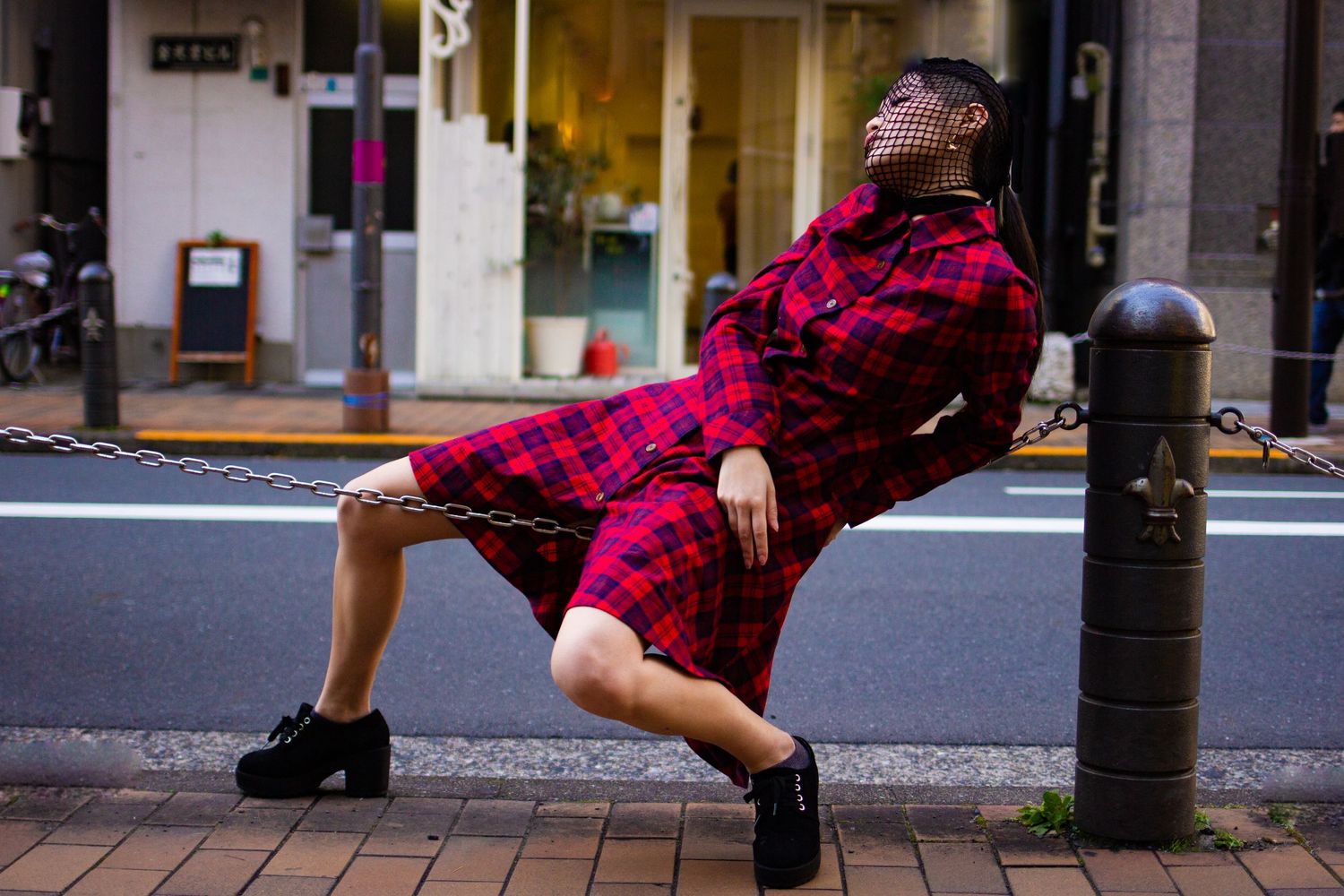 Street fashion model in red plaid dress poses dramatically against urban bollard.