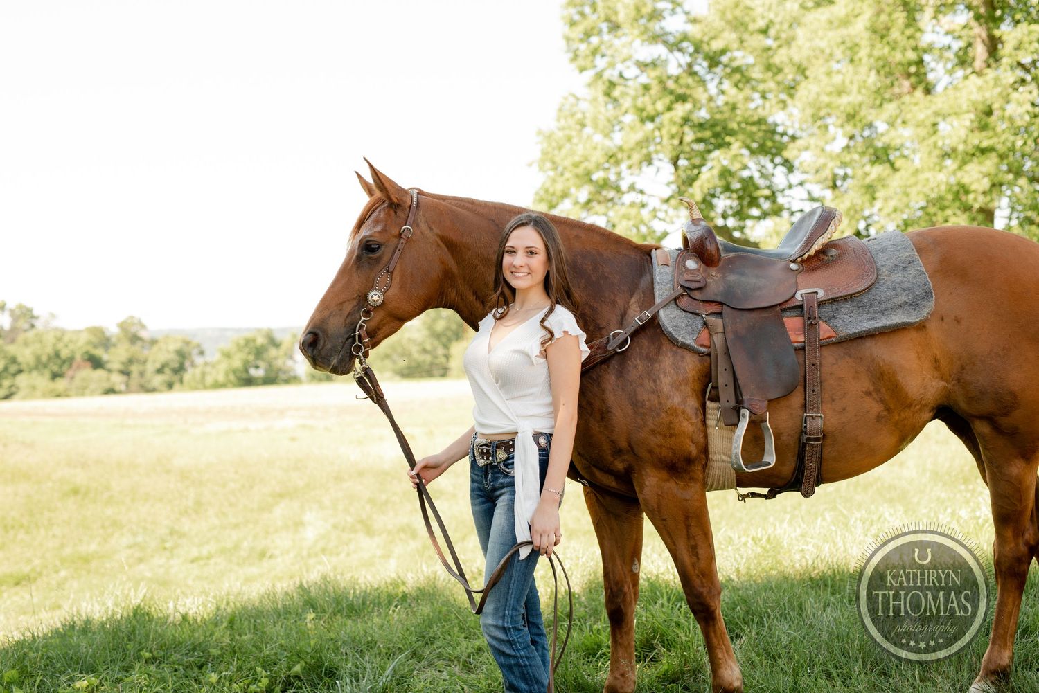 A woman stands with a brown saddled horse in a grassy field on a sunny day.
