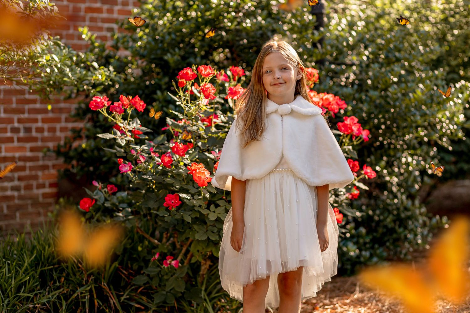 Young girl in white dress and fur cape poses near blooming red roses in garden.