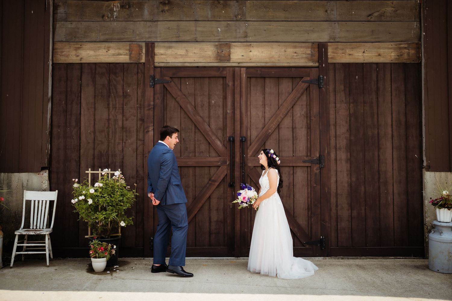 Wedding couple poses playfully in front of rustic wooden barn doors with white chairs and potted plants.