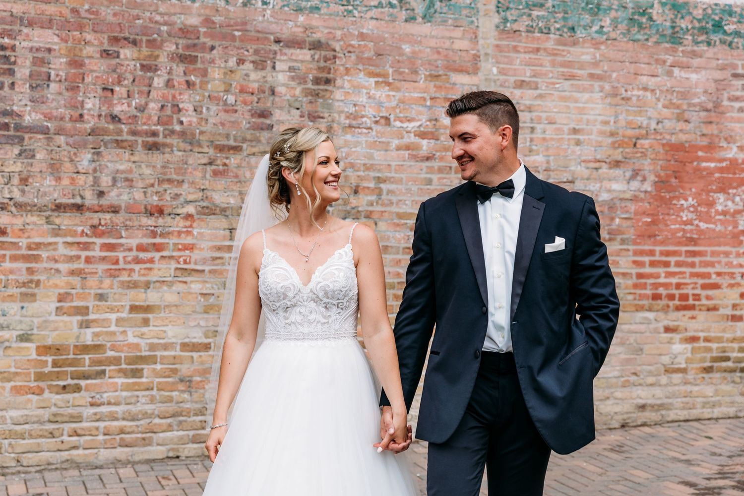 Couple in wedding attire walking together against rustic brick wall backdrop.