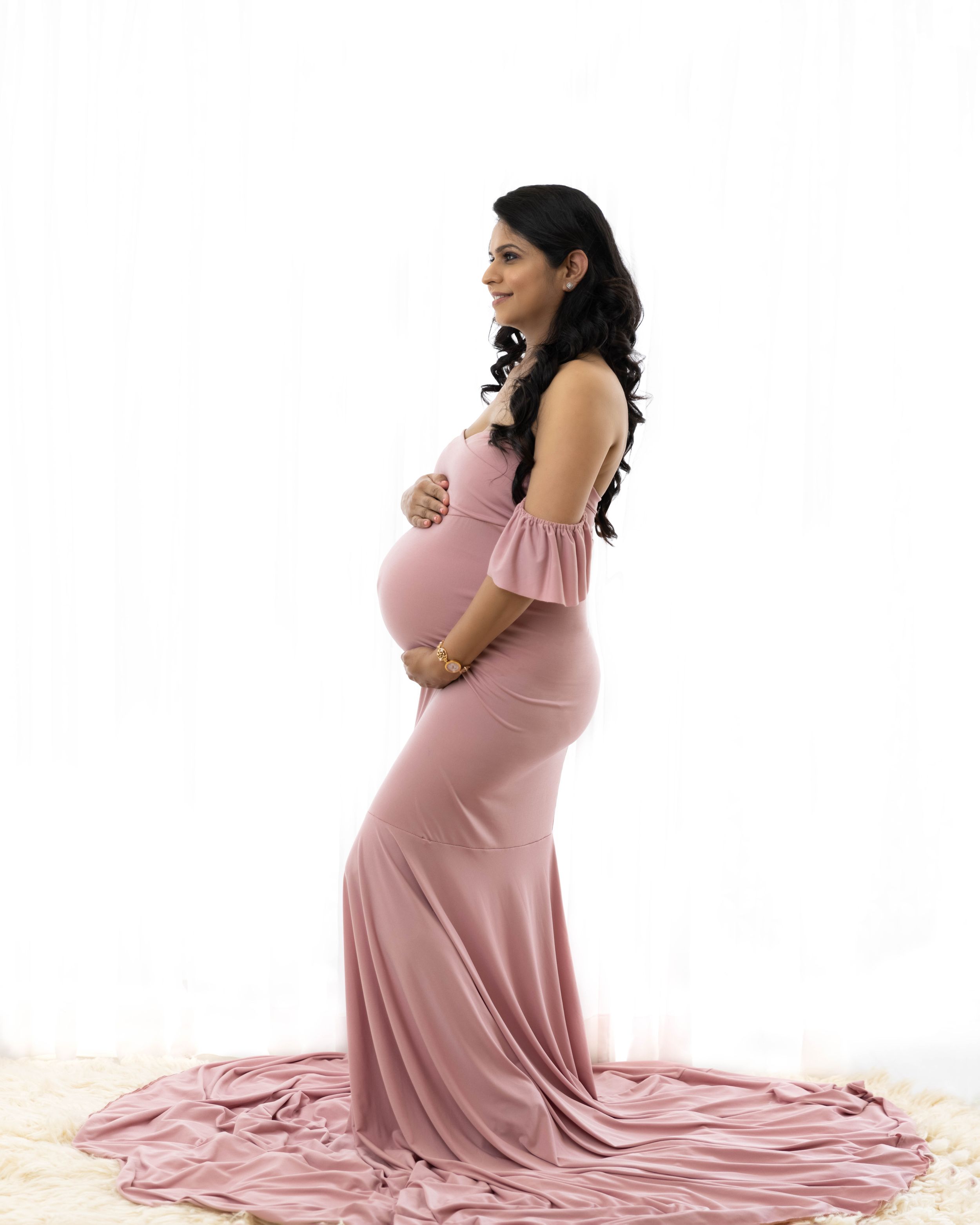 Side view maternity portrait in dusty rose gown with flowing train against white background.