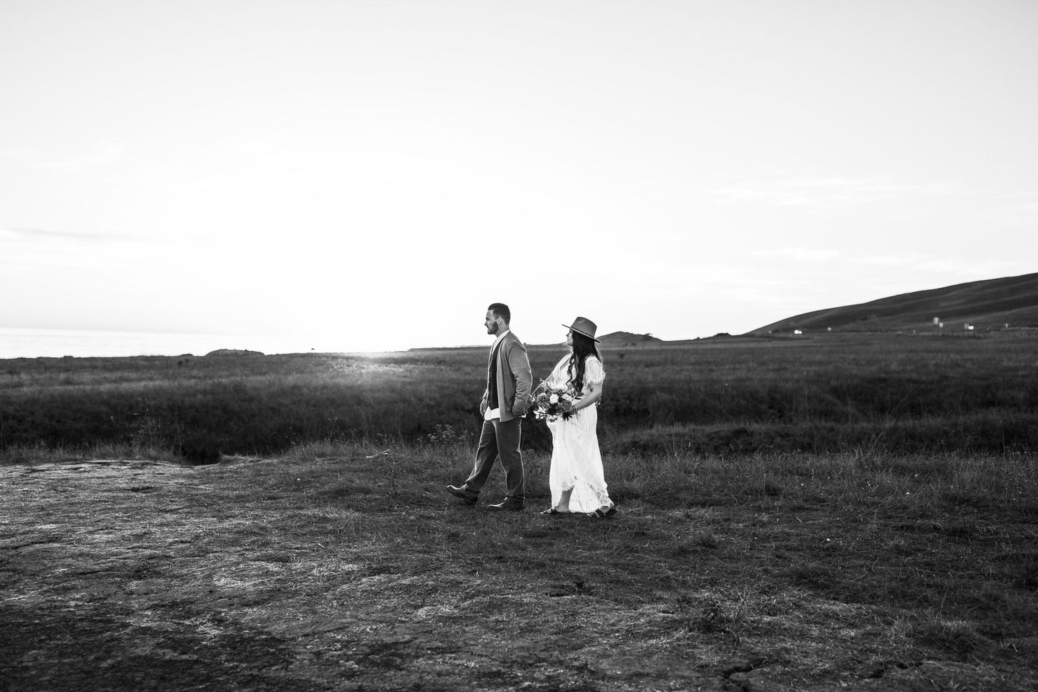 Black and white wedding photo of couple walking through field at sunset.