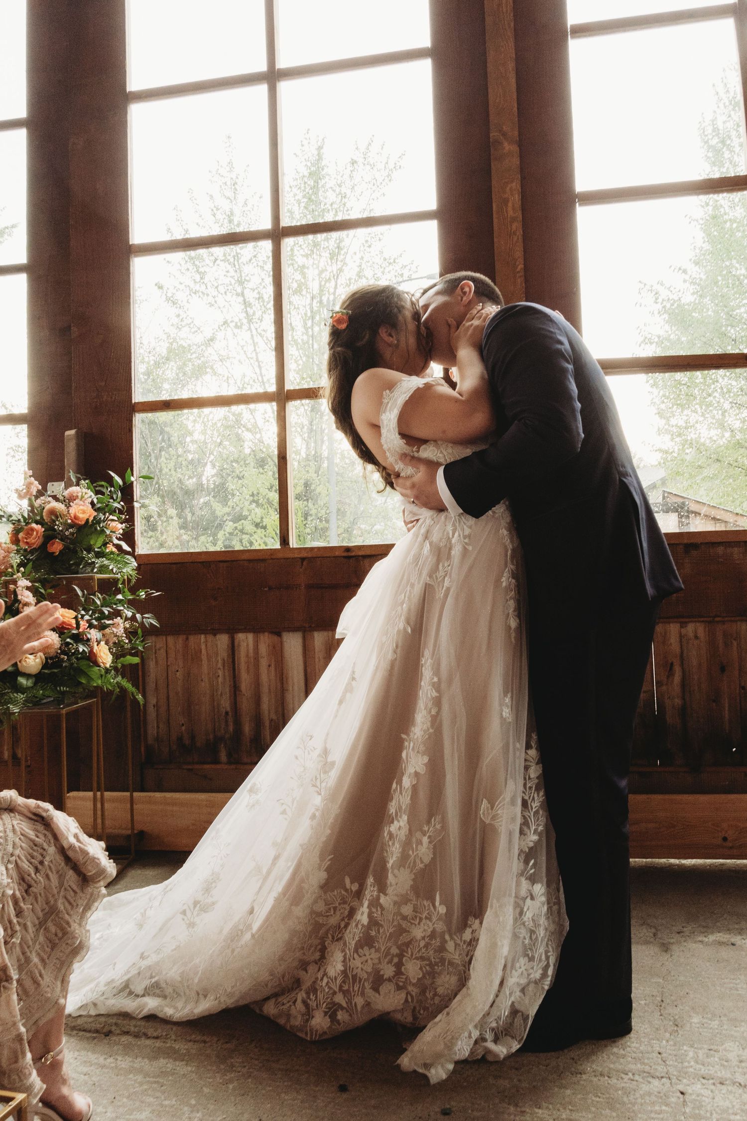A romantic moment as newlyweds share a kiss in front of large windows in a rustic wooden venue with floral decor.
